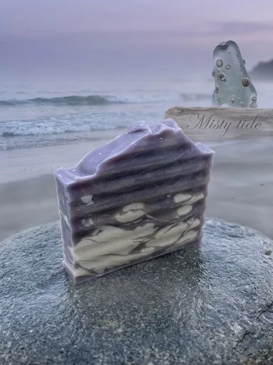 Misty Tide goat milk soap: a layered mauve and cream bar on a stone at dusk, with driftwood and sea glass behind it.