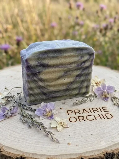 Prairie Orchid goat milk soap: a violet and cream swirled bar on a wood round, surrounded by orchid blossoms and dried grasses.